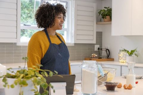 African american home baker smiling while preparing batter with tablet in bright kitchen