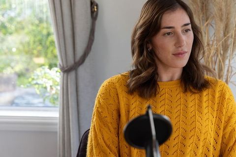 Woman recording podcast at home wearing mustard sweater by window light and dried grasses