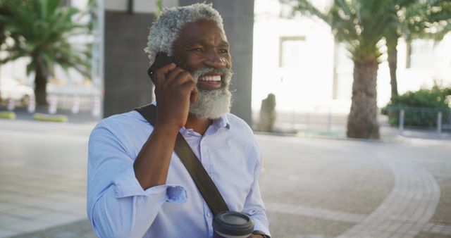 Mature man with a gray beard and casual attire talking on a mobile phone and holding a coffee cup outdoors. Captured in an urban environment with palm trees in the background. Perfect for themes related to communication, modern lifestyle, business, and senior well-being.