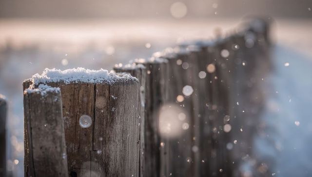 Snow-dusted wooden fence post glowing with golden bokeh and falling snow at sunrise