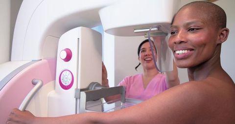 Black woman undergoing mammogram while asian technologist positioning unit, both smiling