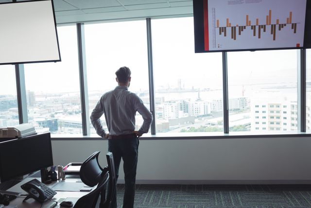 Businessman standing with hands on hips, looking out of office window at cityscape. Ideal for use in business, corporate, and professional contexts, illustrating themes of planning, strategy, and contemplation in a modern workplace.