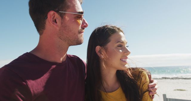 Young couple enjoying a sunny day at the beach, both smiling and looking into the distance. Ideal for use in travel brochures, romantic getaway ads, or summer lifestyle campaigns.