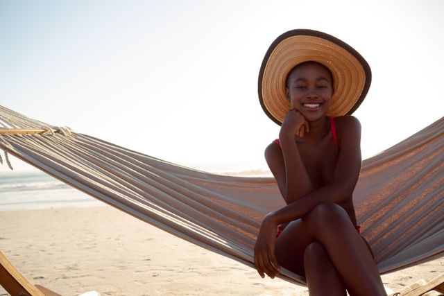 Young woman enjoying a sunny day on the beach while relaxing in a hammock. She is smiling and wearing a wide-brimmed hat, embodying a carefree and happy lifestyle. Perfect for use in travel brochures, summer vacation promotions, lifestyle blogs, and advertisements for beachwear or holiday destinations.