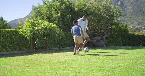 African american father and son practicing soccer dribbling on sunlit backyard lawn