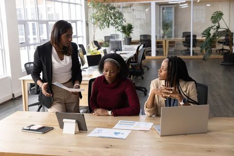 Diverse women collaborating around laptop and tablet in modern sunlit open office planning