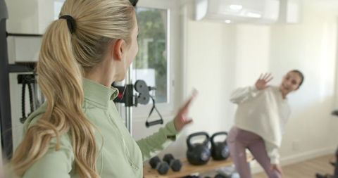 Female friends exercising at home gym waving during kettlebell and dumbbell training