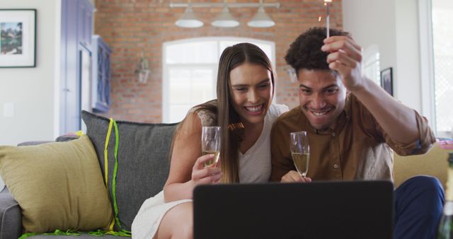 Happy biracial couple drinking champagne at home making celebration laptop image call and laughing. celebration, festivity and communication technology at home.
