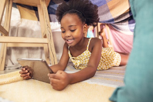 Smiling african american girl with afro hair using digital tablet while lying under blanket tent. Unaltered, wireless technology, childhood, bedding, lifestyle and home concept.