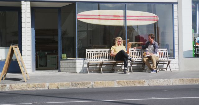 Two friends sitting on a bench outside a surf shop, enjoying a sunny day and engaging in conversation. Great for lifestyle articles, coastal town promotions, surf culture content, and active outdoor lifestyle advertisements.