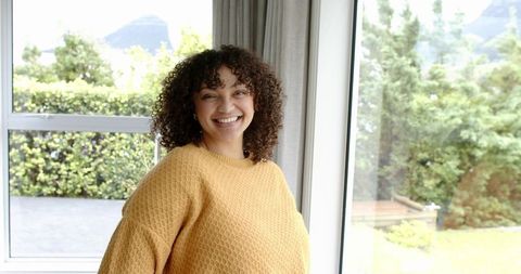 Smiling woman in mustard sweater by large windows enjoying natural light and garden view