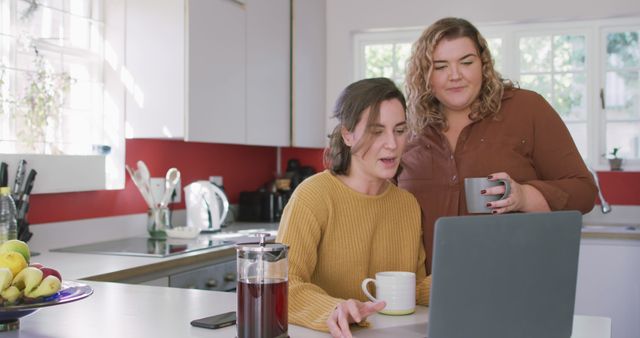 Happy caucasian lesbian drinking coffee and using laptop in kitchen - Download Free Stock Photos Pikwizard.com