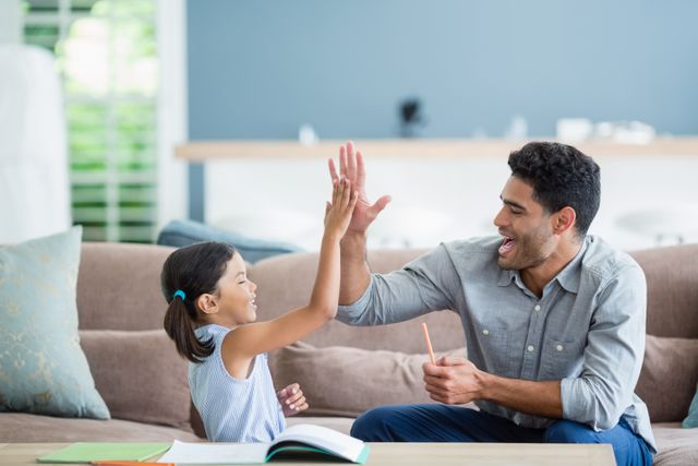 Father and daughter sharing a joyful moment while working on homework together in a cozy living room. Ideal for use in articles or advertisements about family bonding, educational support, parenting tips, and home learning environments.
