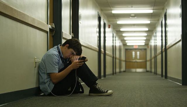 Teen sitting in empty school corridor scrolling smartphone wearing wired headphones