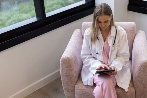 Female clinician sitting by window using tablet in calm clinic interior for telehealth and charting