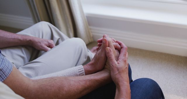 This touching photograph captures an elderly couple seated close together, holding hands affectionately near a sunlit window. Ideal for themes of elderly care, aging with grace, love and companionship in later years, and emotional support. Perfect for use in healthcare, retirement homes, and family-oriented projects emphasizing warmth and connection among seniors.