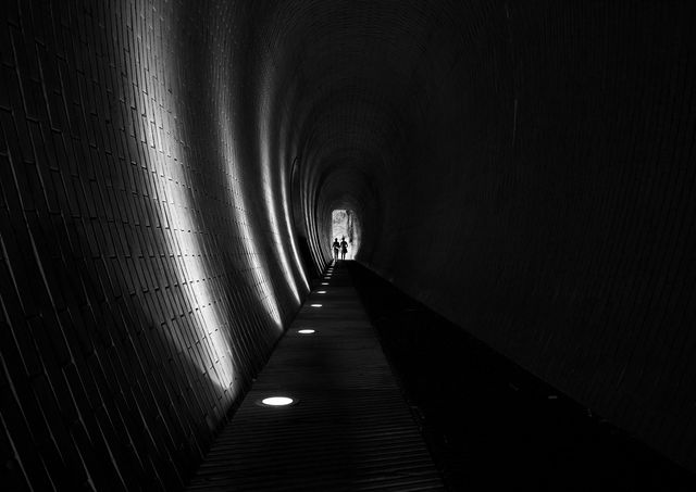 Silhouettes of individuals walking through a long, dark tunnel with circular lights embedded in the floor and a light source at the end, creates dramatic perspective effect. Perfect for use in urban exploration, themes of hope, persistence, and mystery in illustrations, marketing, or editorial content.