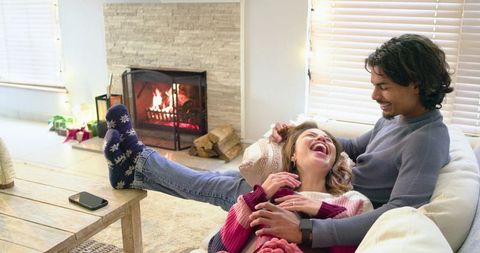 Diverse couple laughing and reclining on couch by fireplace in cozy holiday living room
