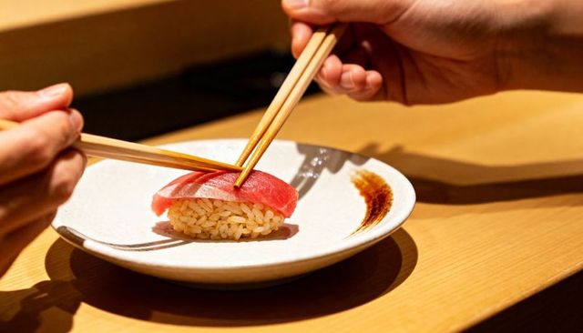 Two hands grasping tuna nigiri with chopsticks at minimal sushi bar on wooden counter