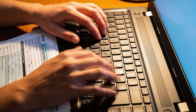 Hands typing on laptop keyboard with form and blue folder on wooden desk, closeup