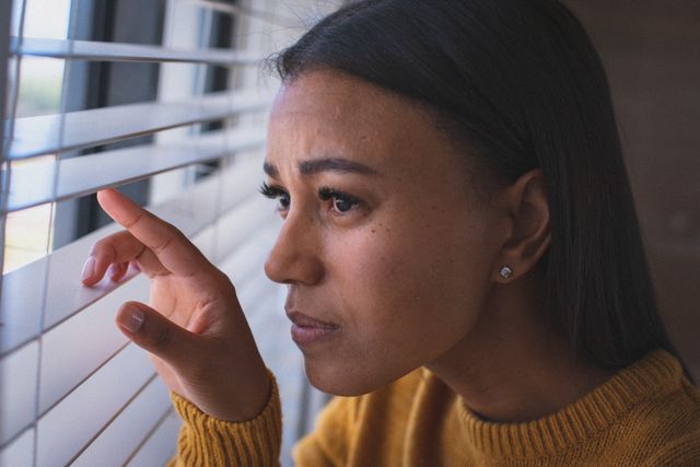This image depicts a woman peering through venetian blinds, looking out of a window. She appears thoughtful and worried, suggesting themes of isolation and contemplation during quarantine. This image can be used in articles or advertisements related to mental health, quarantine experiences, or the impact of lockdowns on daily life.