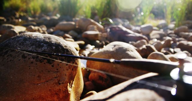 A fishing rod rests on a bed of smooth river stones bathed in warm sunlight, suggesting a peaceful fishing trip. The scene evokes a sense of relaxation and connection with nature.