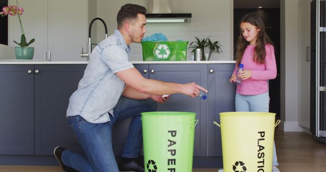 Father and daughter are sorting recyclables in a modern kitchen. The father is squatting next to two recycling bins labeled 'Paper' and 'Plastic' while the daughter holds a plastic bottle. Ideal for use in educational materials about recycling, sustainability initiatives, and family-oriented eco-friendly activities.