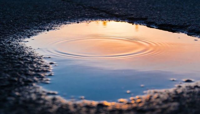 Shallow Puddle Reflecting Orange-Blue Sunset with Circular Ripples on Gritty Asphalt