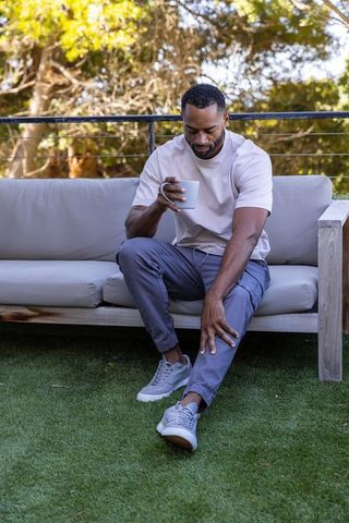 African american man relaxing on backyard sofa holding coffee mug reaching leg on sunlit patio
