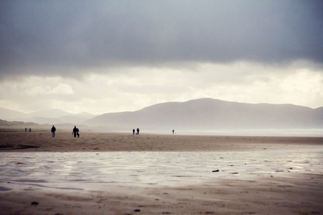 Few people are seen strolling on an expansive sandy beach under a cloudy, dramatic sky with distant mountains. This serene coastal scene is suitable for use in travel brochures, nature blogs, and relaxation-themed projects.