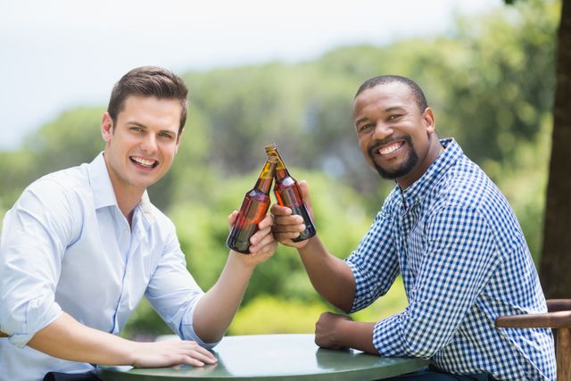 Two friends toasting beer bottles while smiling outdoors. Ideal for use in advertisements promoting social gatherings, restaurants, or beer brands. Perfect for illustrating themes of friendship, celebration, and leisure.