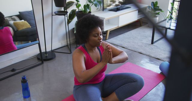 African American woman practicing yoga and meditation at home sitting on a pink yoga mat. Surrounded by indoor plants, she creates a peaceful and serene atmosphere. She holds her hands in a prayer position, focusing on inner peace and mindfulness. This image can be used for promoting home wellness, fitness routines, mental health, and yoga practice.