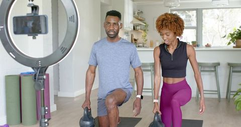 Diverse couple performing kettlebell lunges in modern kitchen home workout with ring light