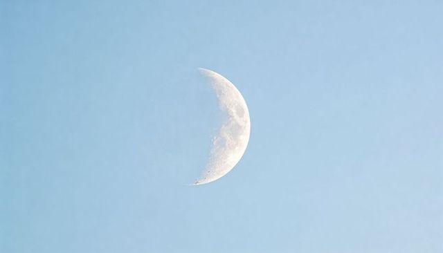 Waning crescent moon revealing cratered surface along terminator against pale blue sky