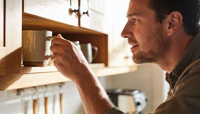 Man reaching for ceramic mug in sunlit kitchen cupboard morning routine