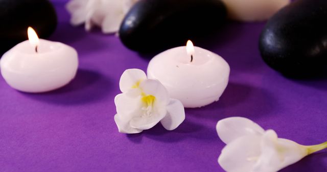 Lit candles and smooth stones are arranged on a purple surface, creating a tranquil spa atmosphere. The white flowers add a touch of natural beauty, enhancing the calming effect of the scene.
