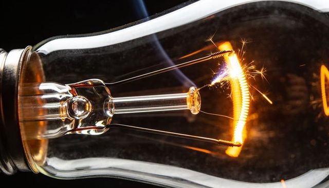 Glowing incandescent bulb macro showing coiled filament sparking over glass stem warm amber
