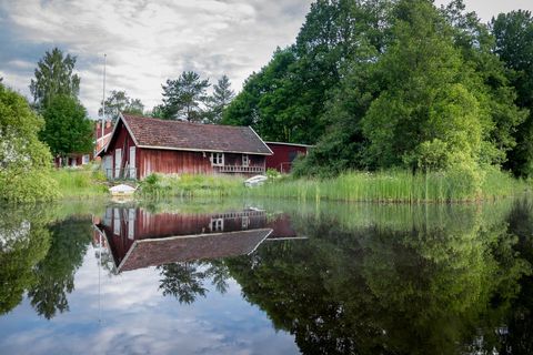 Red boathouse reflecting on calm Scandinavian lake with lush trees and rowboat
