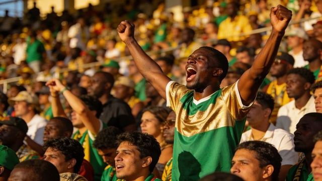 Ecstatic fan cheering, raising arms in sunlit stadium crowd wearing green gold jersey