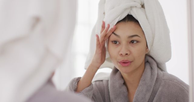 Image of portrait of smiling biracial woman with towel on hair looking in mirror in bathroom. Health and beauty, leisure time, domestic life and lifestyle concept.
