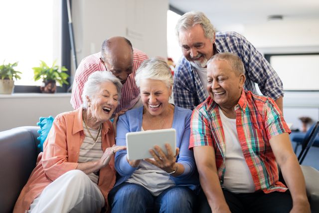 Group of cheerful senior friends gathered around a digital tablet, enjoying each other's company in a nursing home. Perfect for illustrating themes of technology use among the elderly, social bonding in retirement, and the joy of togetherness in later life. Ideal for use in advertisements, articles, and promotional materials related to senior living, healthcare, and technology for seniors.