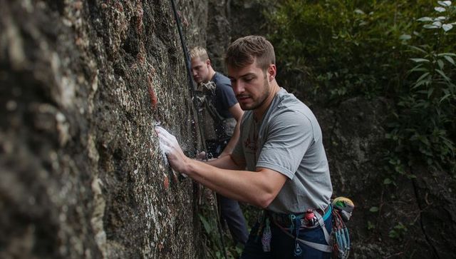 Rock climber preparing chalked hands on forest cliff with harness and rope, teamwork focus