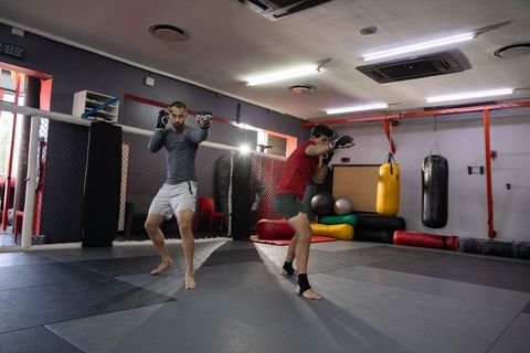 Two mma fighters sparring on mat inside martial arts gym with bags and cage