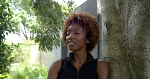Serene african american woman leaning on tree in lush courtyard wearing black zip top