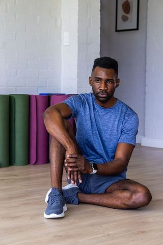 African man sitting on studio floor near colorful yoga mats wearing blue activewear