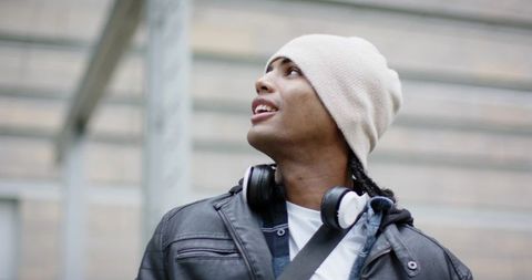 African American man looking up on city street wearing beanie, headphones, leather jacket