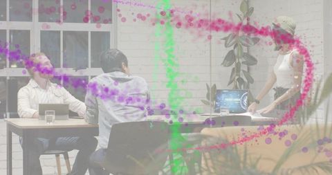 Woman in beanie presenting to team at collaborative office table with laptops and plants