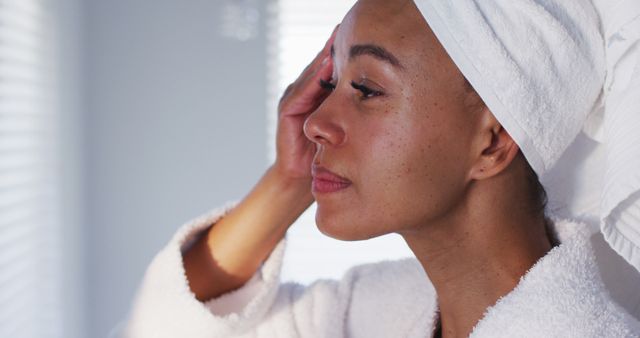 Woman practicing daily skincare routine, applying lotion or cream while wearing a white bathrobe and towel around her hair. Appears composed and focused, highlighting dedicated personal grooming and self-care habits. Perfect for advertisements promoting skincare products, beauty and wellness blogs, or spa and self-care promotions.