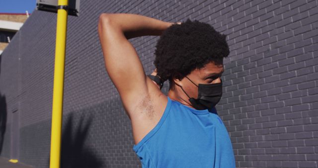 African American man stretching his arm while wearing a face mask outdoors. He is getting ready for exercising in an urban environment with a brick wall in the background. Ideal for content related to fitness, urban lifestyle, pandemic health precautions, or outdoor activities.