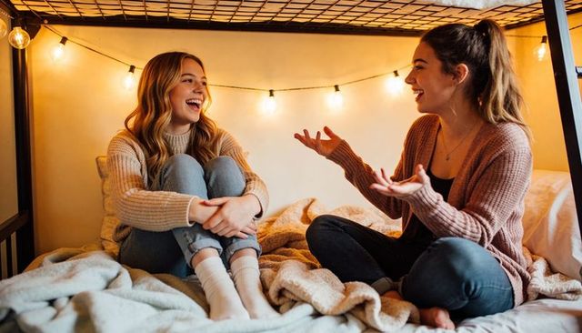 College roommates laughing on lower bunk bed with warm string lights and cozy blankets
